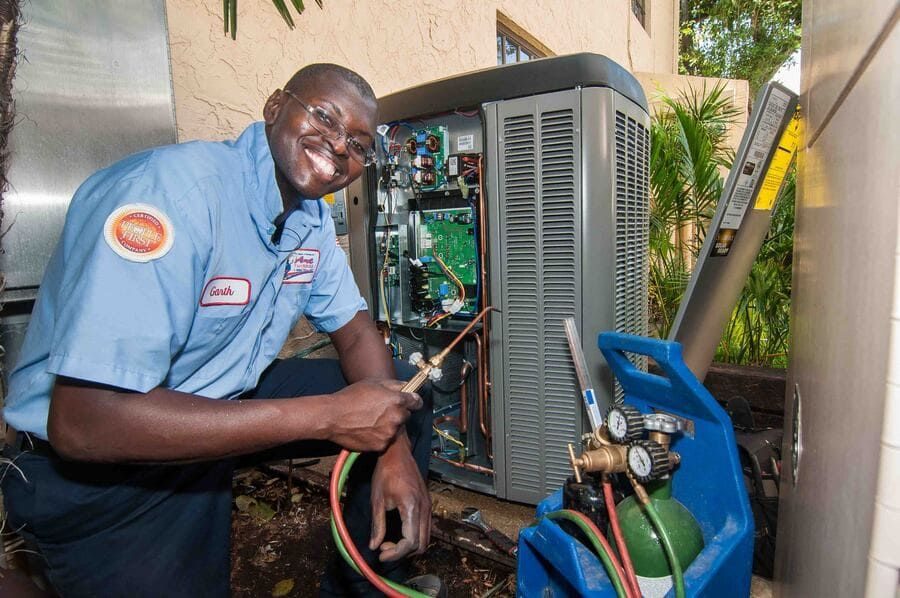 HVAC technician in a blue uniform crouches beside an open outdoor air conditioner unit, using hoses and gauges to repair it.